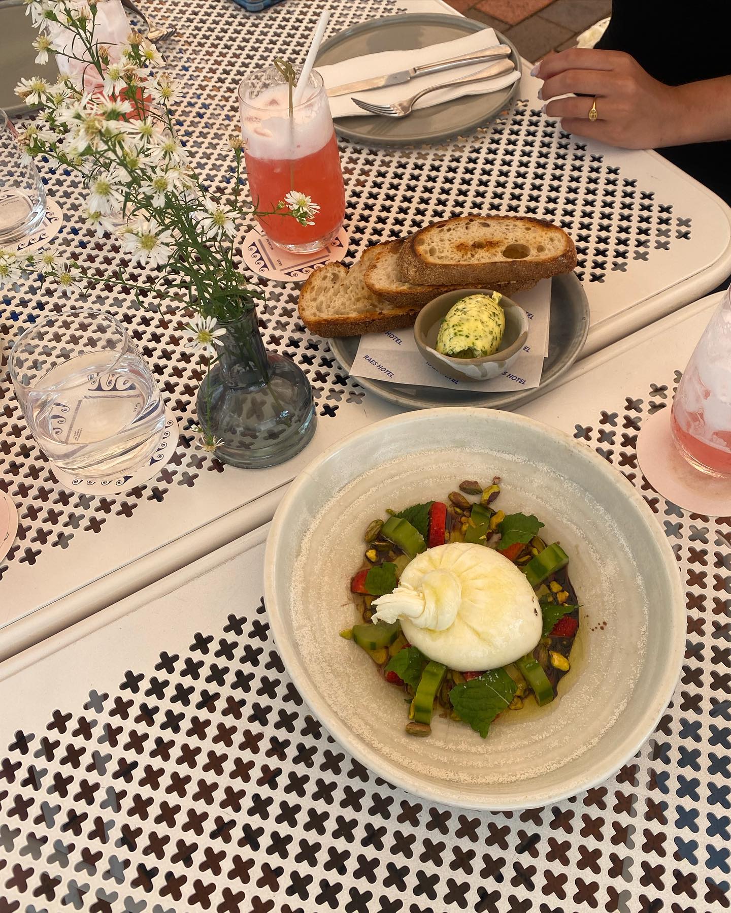 Overhead shot of burrata salad, bread, cocktail, and flowers on a white table, showcasing fresh food and drink in a casual setting.