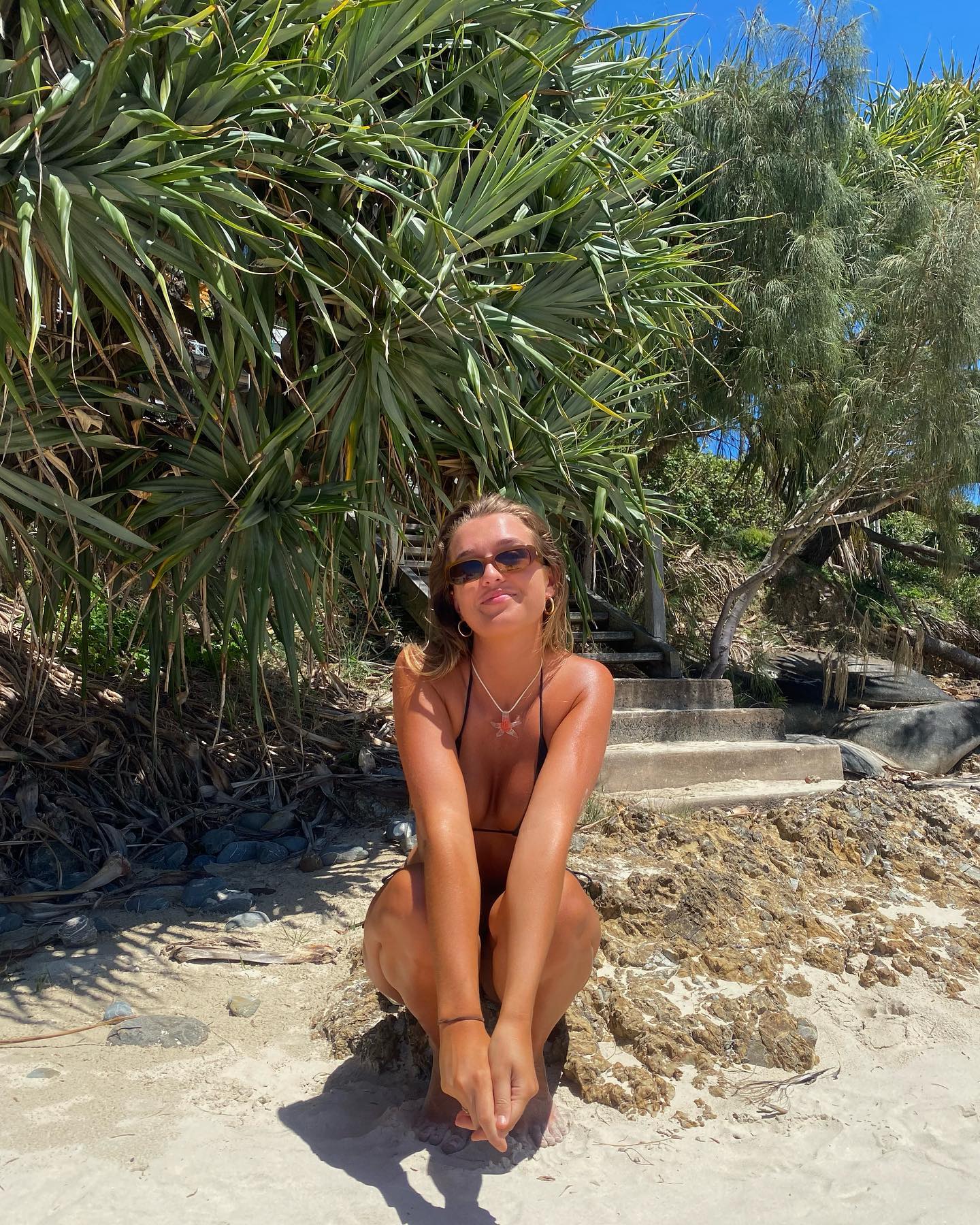Blonde woman in bikini sits on beach rocks surrounded by green foliage on a sunny day with a blue sky in the background.