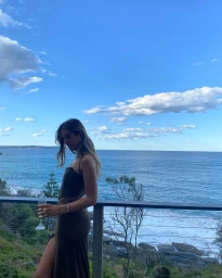 Woman in dark sleeveless dress holding a glass on a balcony overlooking the ocean in Sydney by Kate Robinson