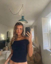 A young woman taking a mirror selfie in a living room, wearing an off-the-shoulder dark top and holding a smartphone.