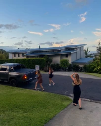 Three women walking on an asphalt street near a dark pickup truck and a two-story residential building under a cloudy sky.