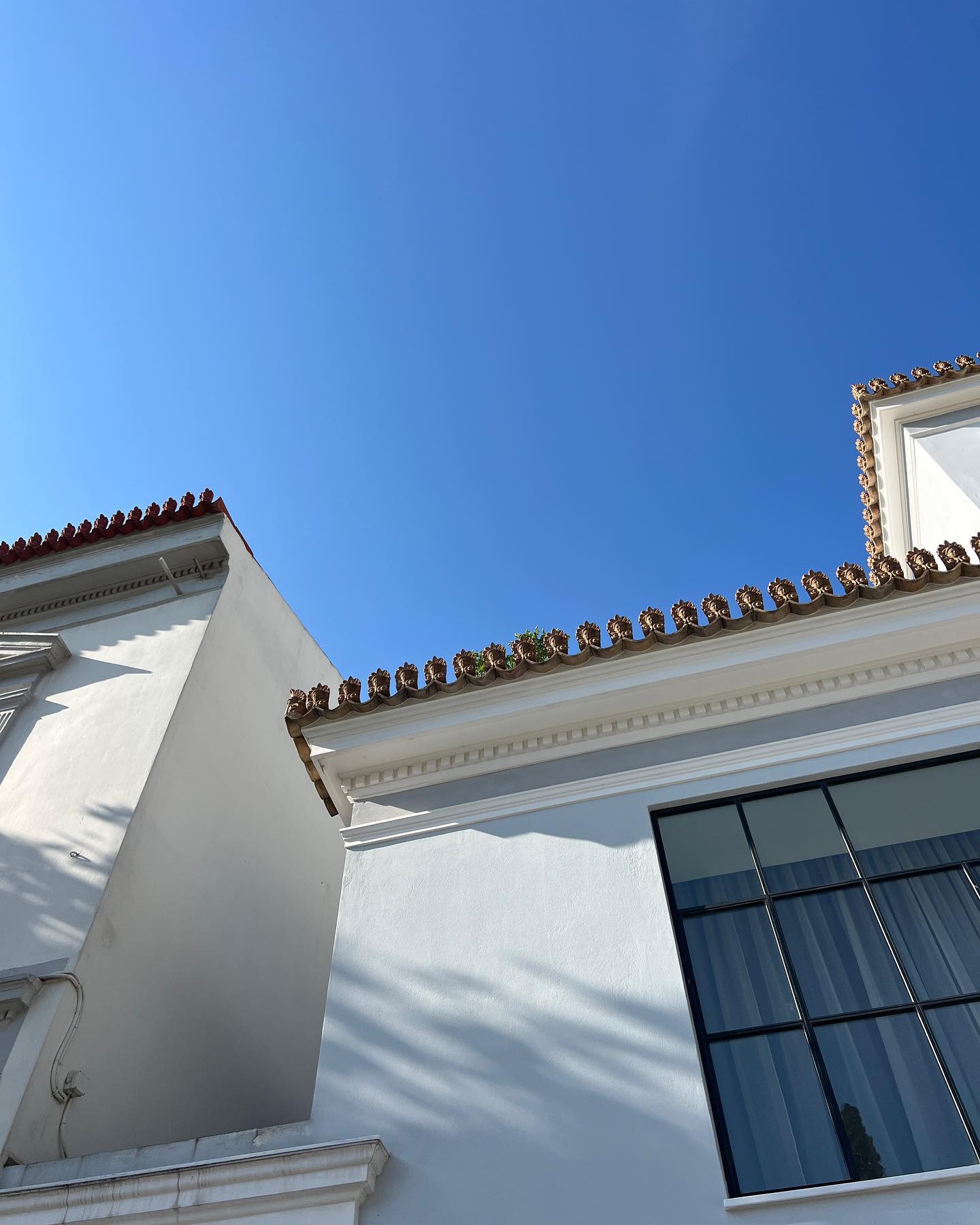 Exterior of a white building with a window under a clear blue sky, featuring terracotta roof tiles, architectural details, and strong sunlight.