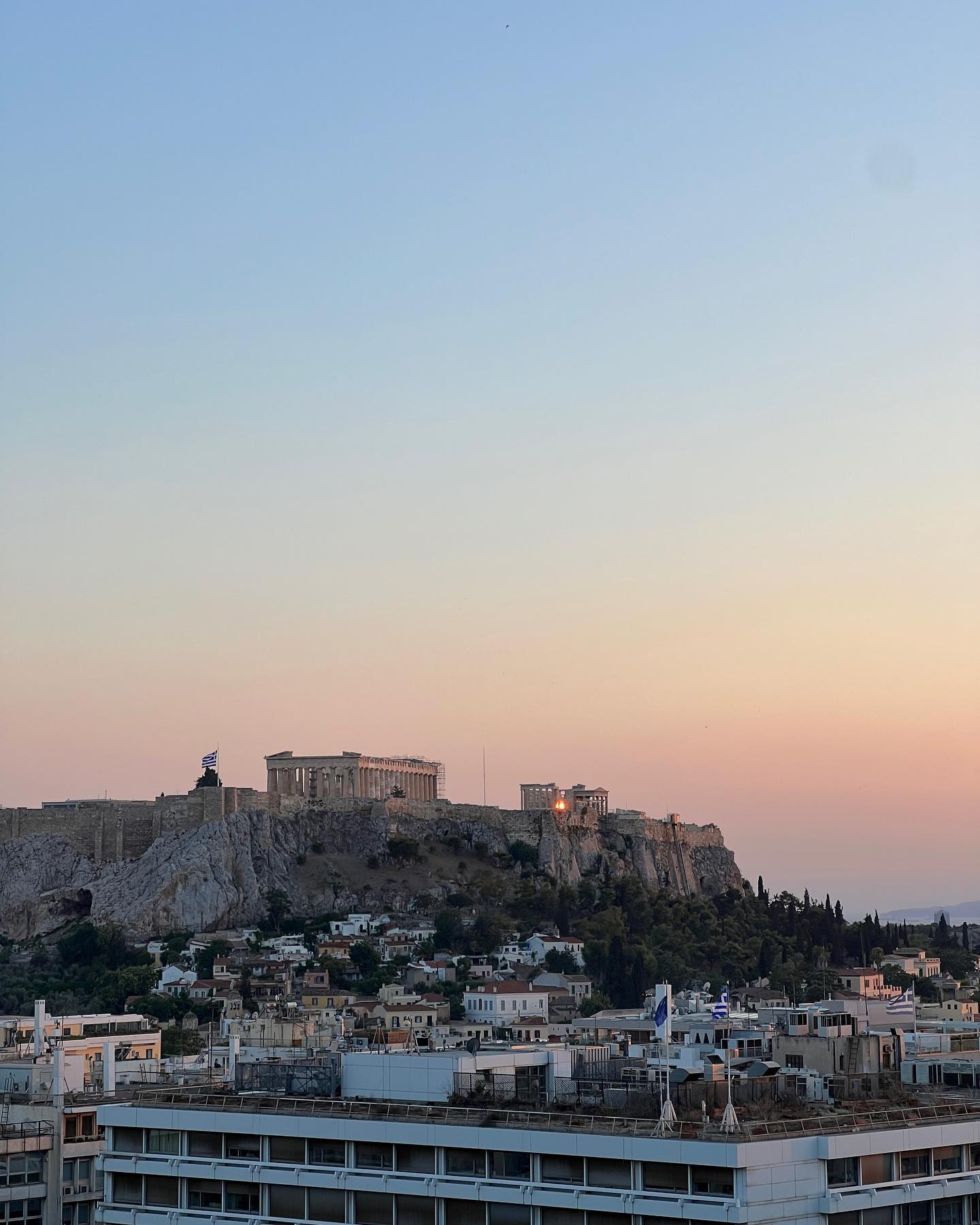 Athens cityscape featuring the Acropolis on a rocky hill during sunset, with city buildings and trees in the foreground.