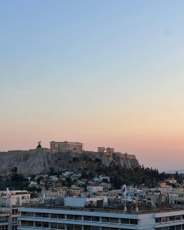 Athens cityscape featuring the Acropolis on a rocky hill during sunset, with city buildings and trees in the foreground.