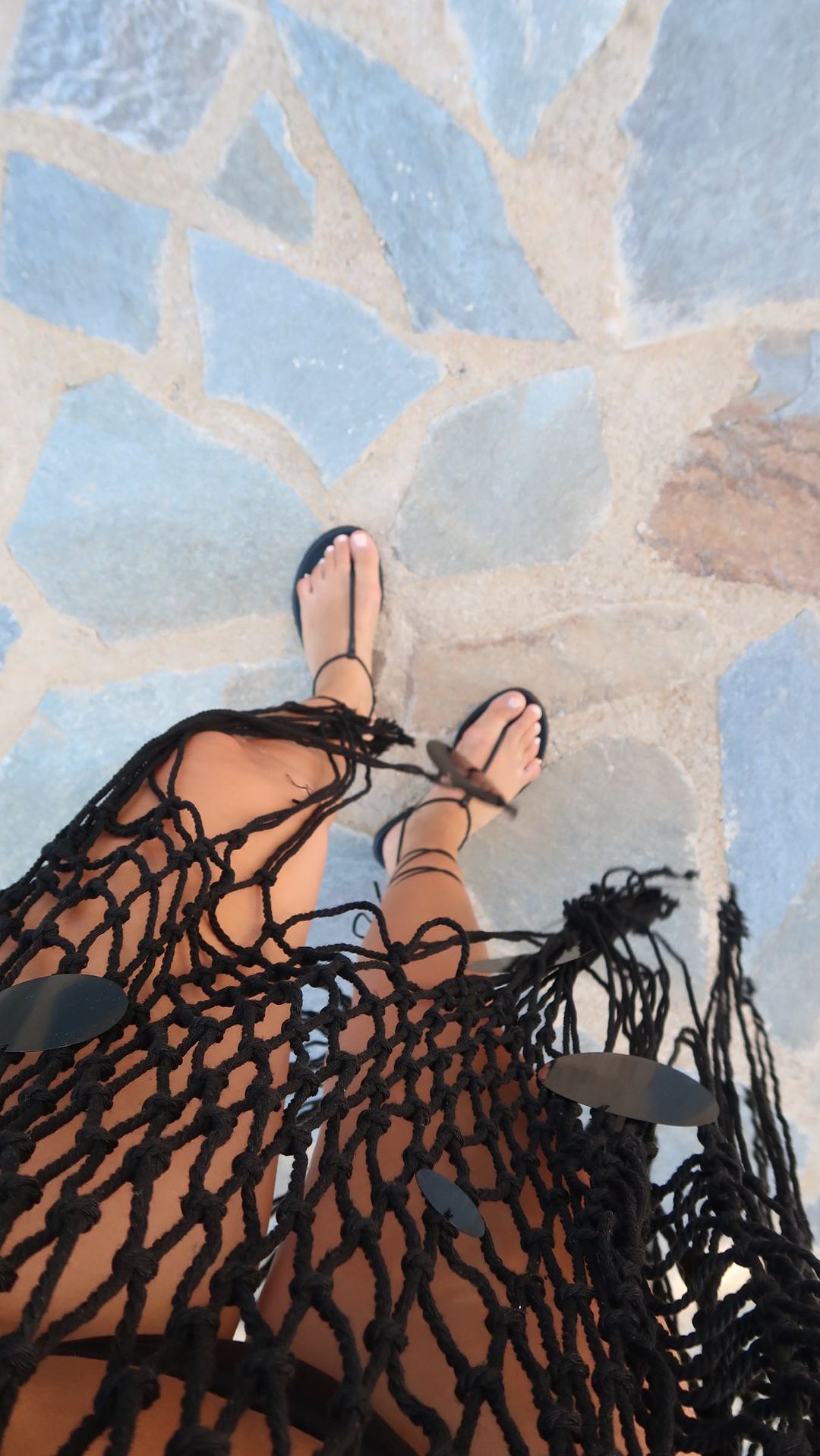 Person wearing black sandals and a woven coverup stands on a stone tile floor in this eye-level shot.