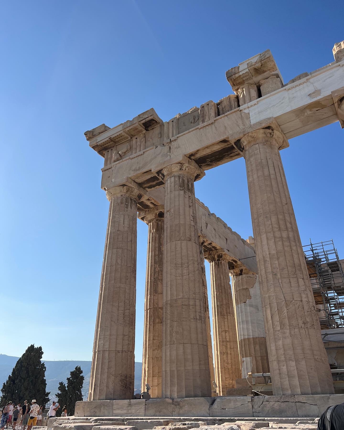 Low-angle view of the Parthenon ruins in Athens, Greece, showcasing its ancient columns against a bright blue sky and scattered greenery.