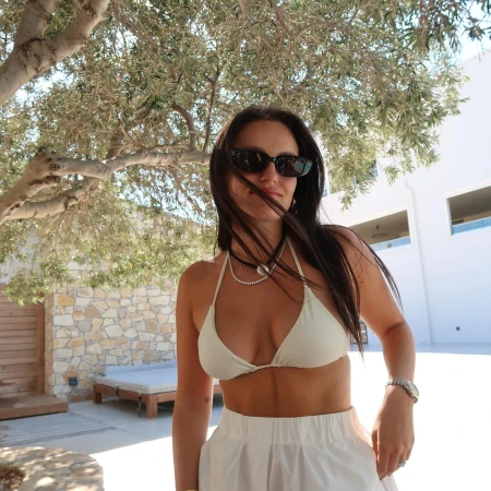 Woman in white bikini top and white skirt wearing sunglasses under a tree by Katherine Denton