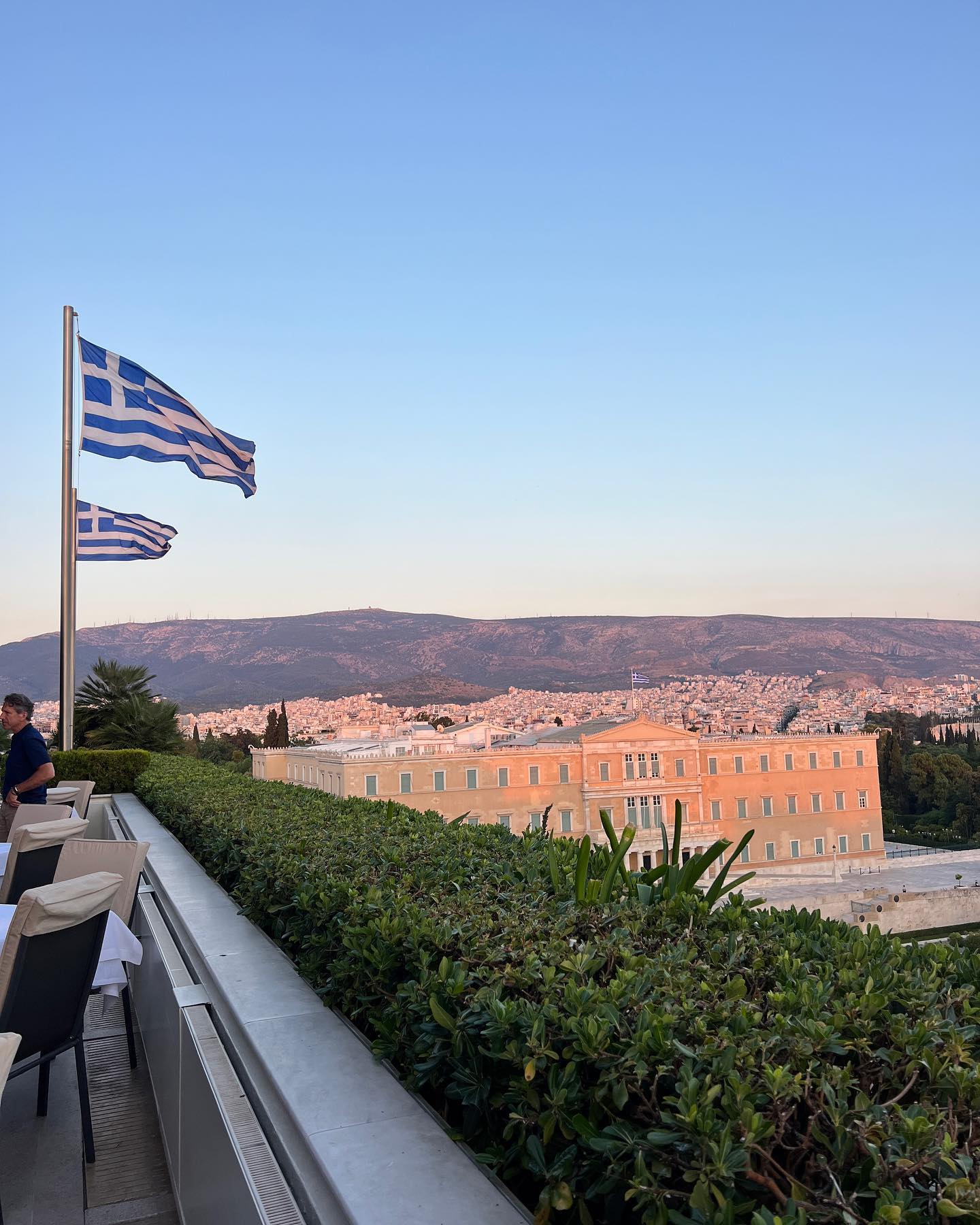 Daytime outdoor shot of two Greek flags, a building complex and landscape of Athens, Greece, featuring city skyline in the background.
