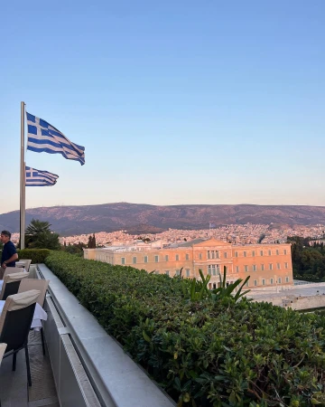 Daytime outdoor shot of two Greek flags, a building complex and landscape of Athens, Greece, featuring city skyline in the background.