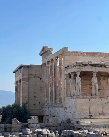 Ancient Greek architecture of the Erechtheion temple, featuring classical columns, statues, and stonework, set against a clear blue sky.