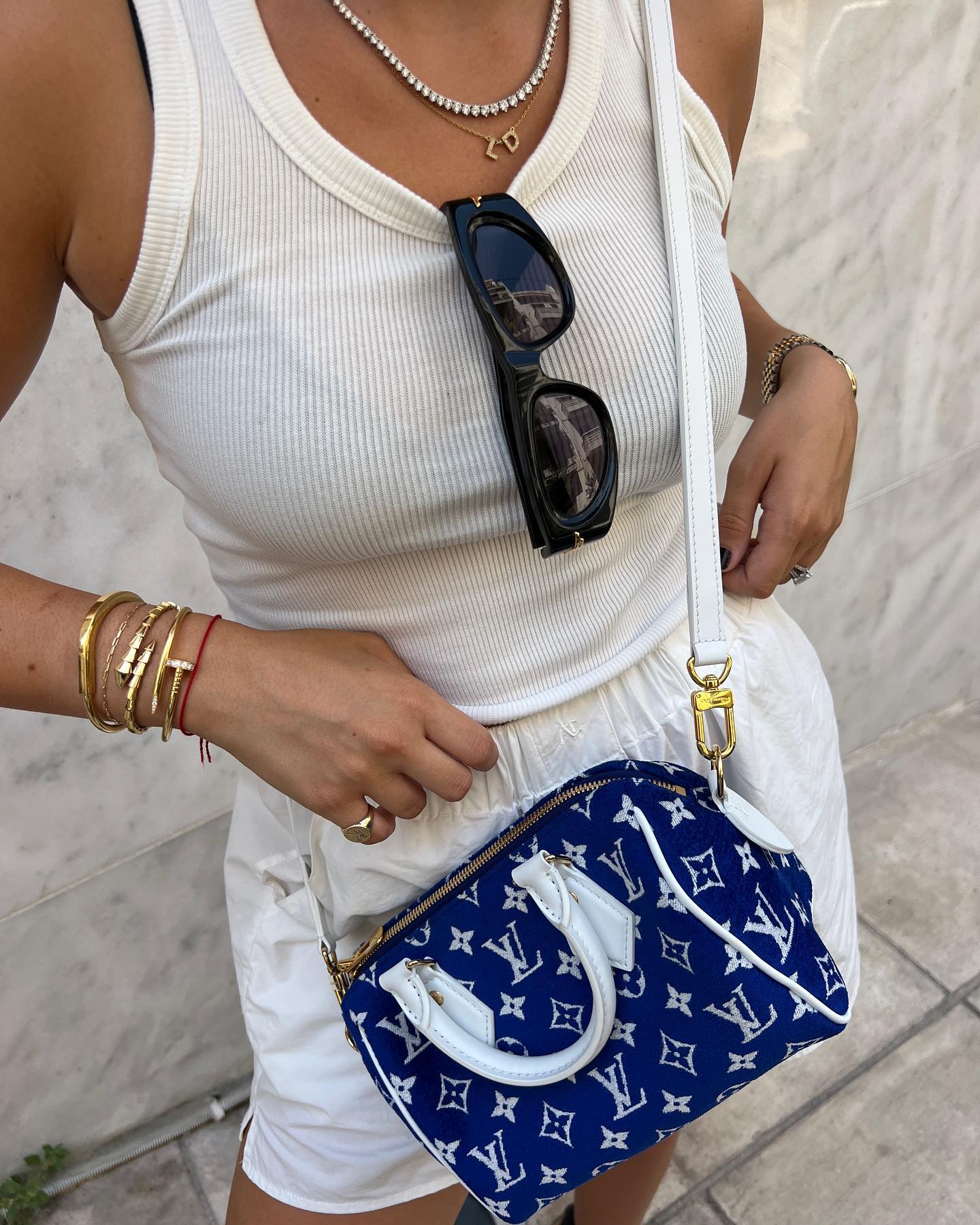 A woman wears a white tank top, shorts, gold jewelry, and a Louis Vuitton handbag, against a neutral background, showcasing fashion and style.