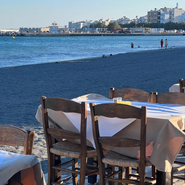 Beachside restaurant table setting with ocean view, featuring dark wooden chairs and a white tablecloth under bright daylight.