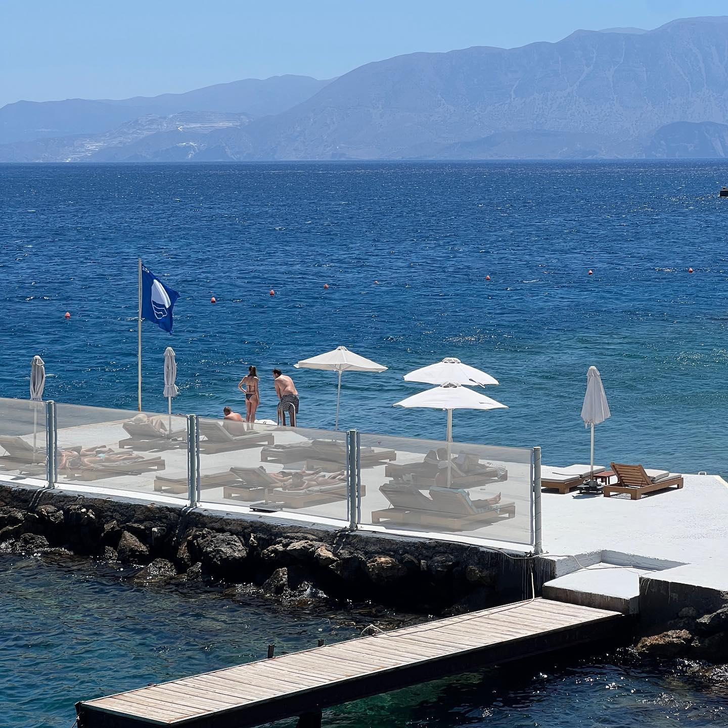 People relaxing on lounge chairs by the sea on a sunny day. Umbrellas provide shade on a stone pier, mountains visible across the water.