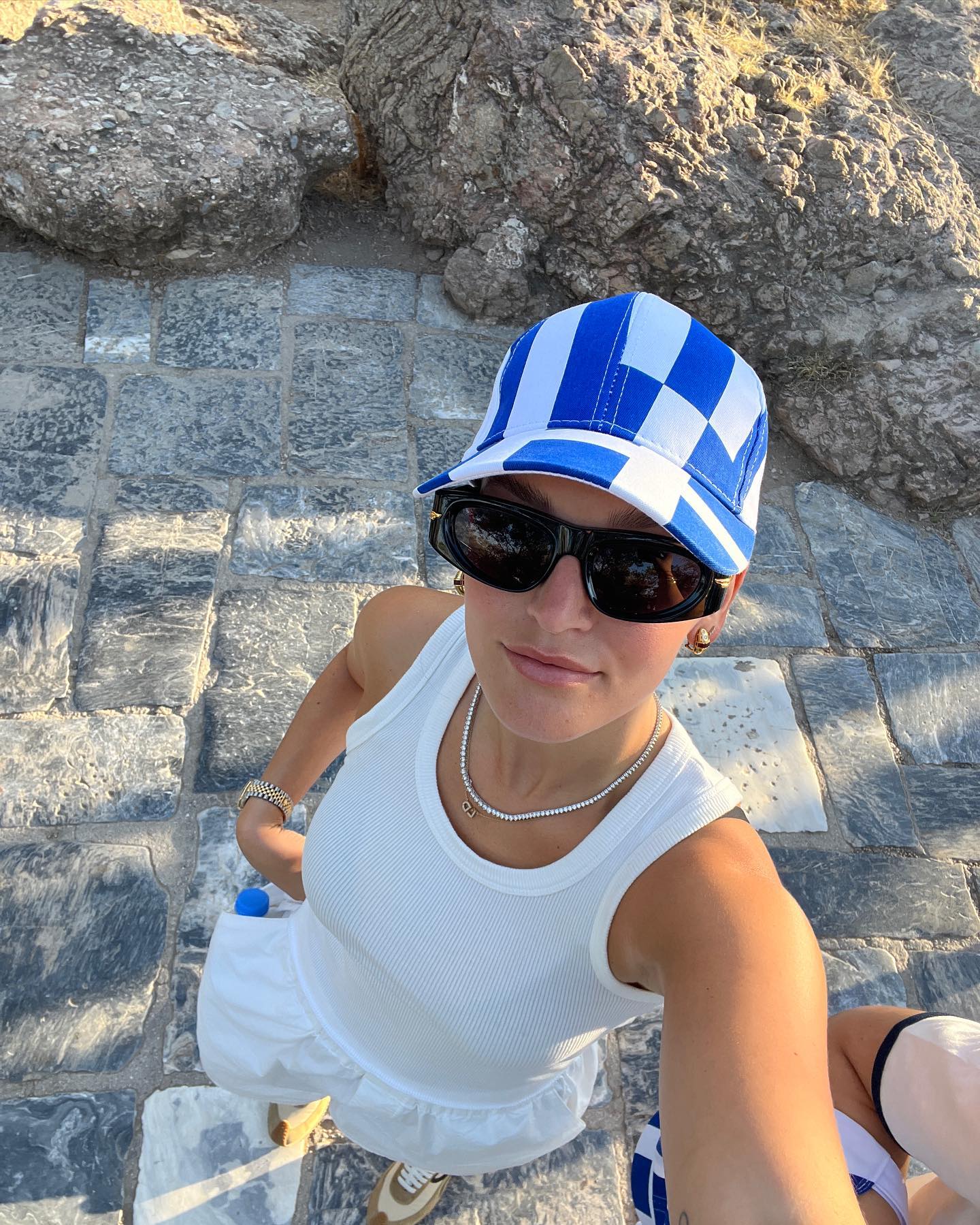 Woman in a white dress and blue and white cap takes a selfie outdoors with a rocky and stone background on a sunny day.