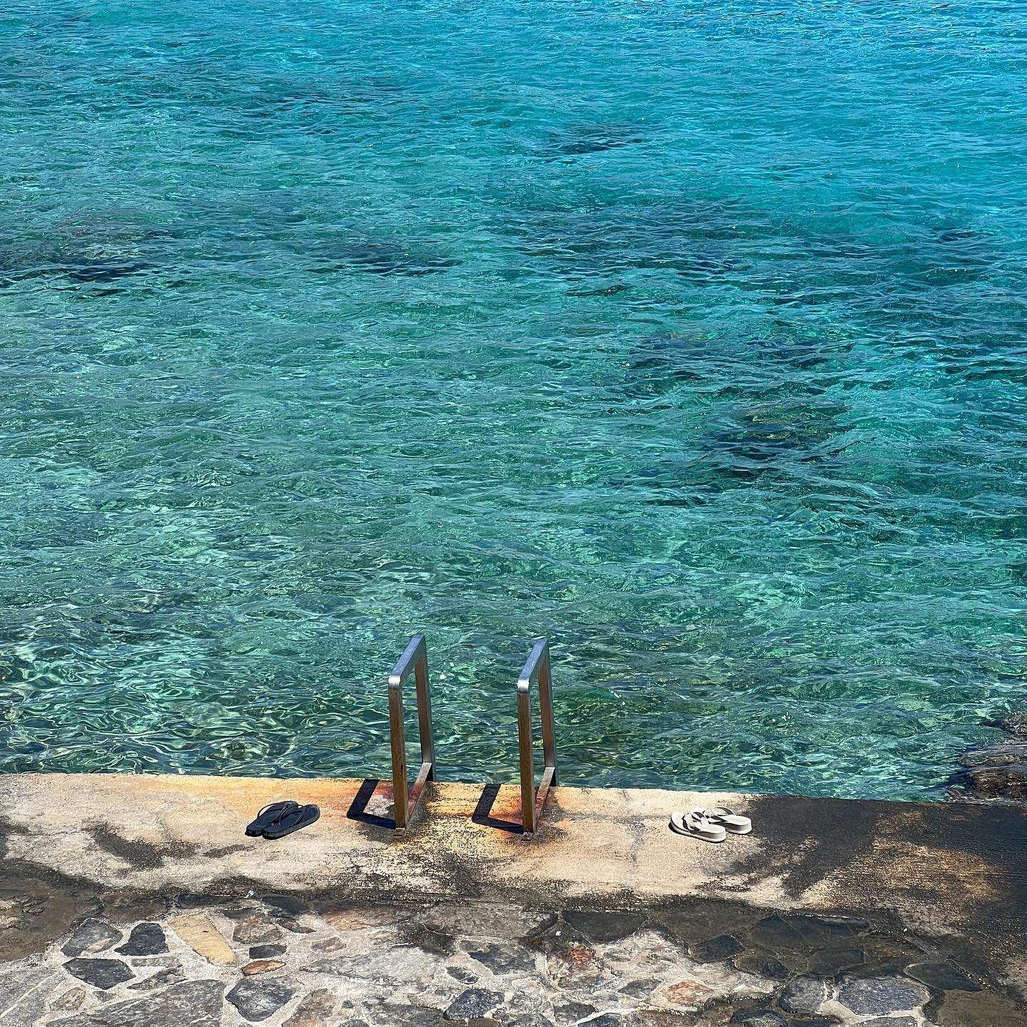 Tranquil turquoise water surrounds concrete platform with ladder and pairs of sandals, creating a peaceful seaside scene on a sunny day.