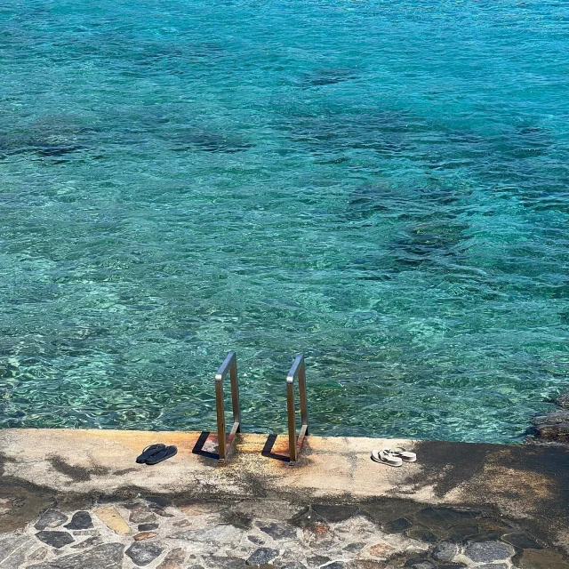 Tranquil turquoise water surrounds concrete platform with ladder and pairs of sandals, creating a peaceful seaside scene on a sunny day.