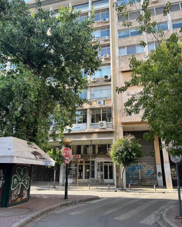 Tall office building with rows of windows and greenery in urban street scene with sidewalks, roads, and a small vendor kiosk.