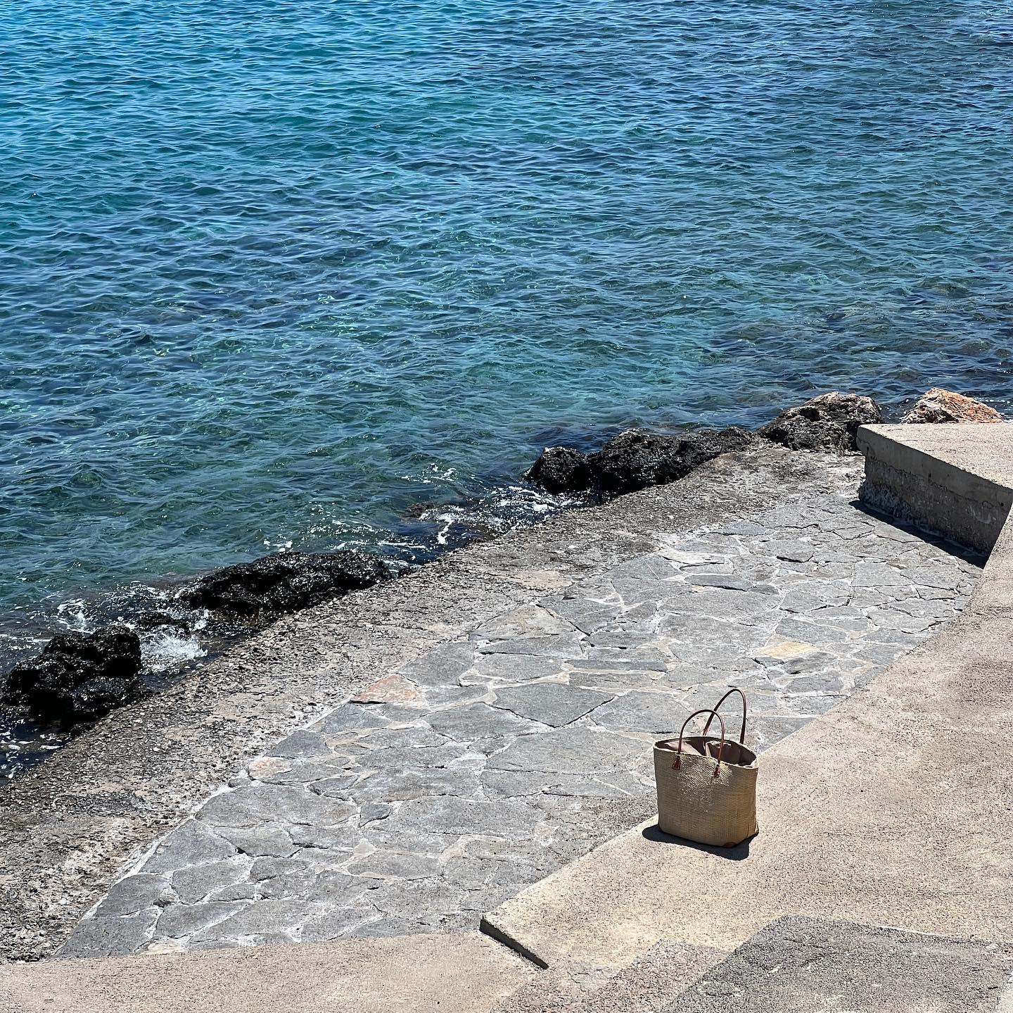 Straw tote bag on a concrete step near the turquoise sea on a sunny day, showcasing a simple design and seaside setting.
