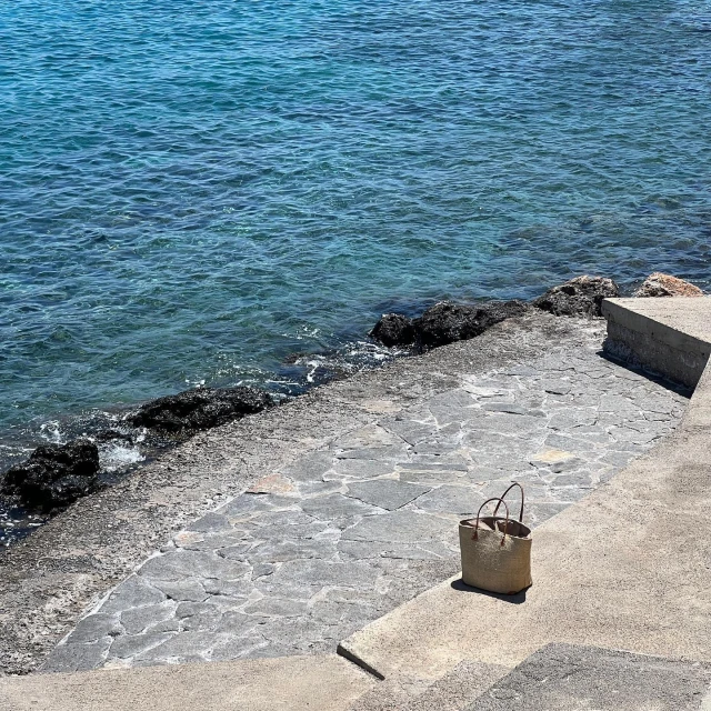 Straw tote bag on a concrete step near the turquoise sea on a sunny day, showcasing a simple design and seaside setting.