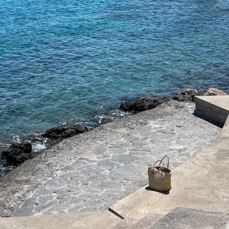 Straw tote bag on a concrete step near the turquoise sea on a sunny day, showcasing a simple design and seaside setting.