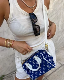 Woman in white ribbed tank top wearing stacked gold bracelets, holding a blue patterned handbag against a stone wall.