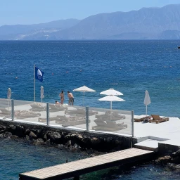 Coastal resort terrace with sun loungers, white umbrellas, and distant mountains across blue water by Katherine Denton