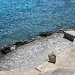 A woven straw tote bag resting on a stone promenade next to clear blue ocean water in Sydney.