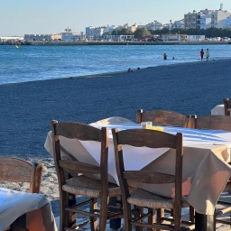 Outdoor restaurant tables overlooking a blue sea with distant town buildings and people walking along the shore.