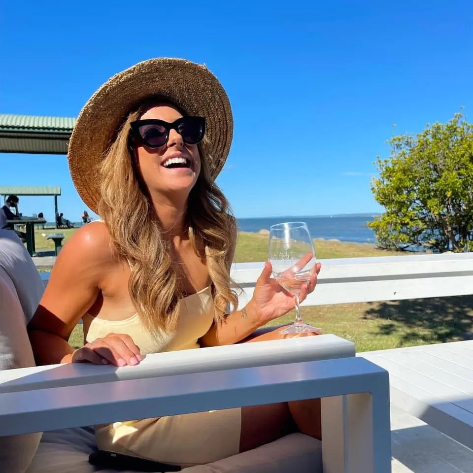 Smiling woman in sunglasses and sun hat sits outside holding a wine glass on a sunny day with a blue sky and trees in the background.