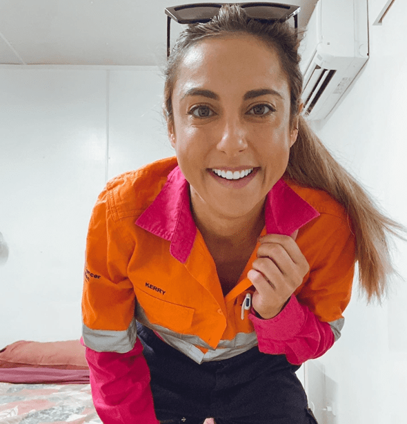 Cheerful woman in orange and pink work jumpsuit smiles indoors, wearing sunglasses on her head and posed in a medium shot.
