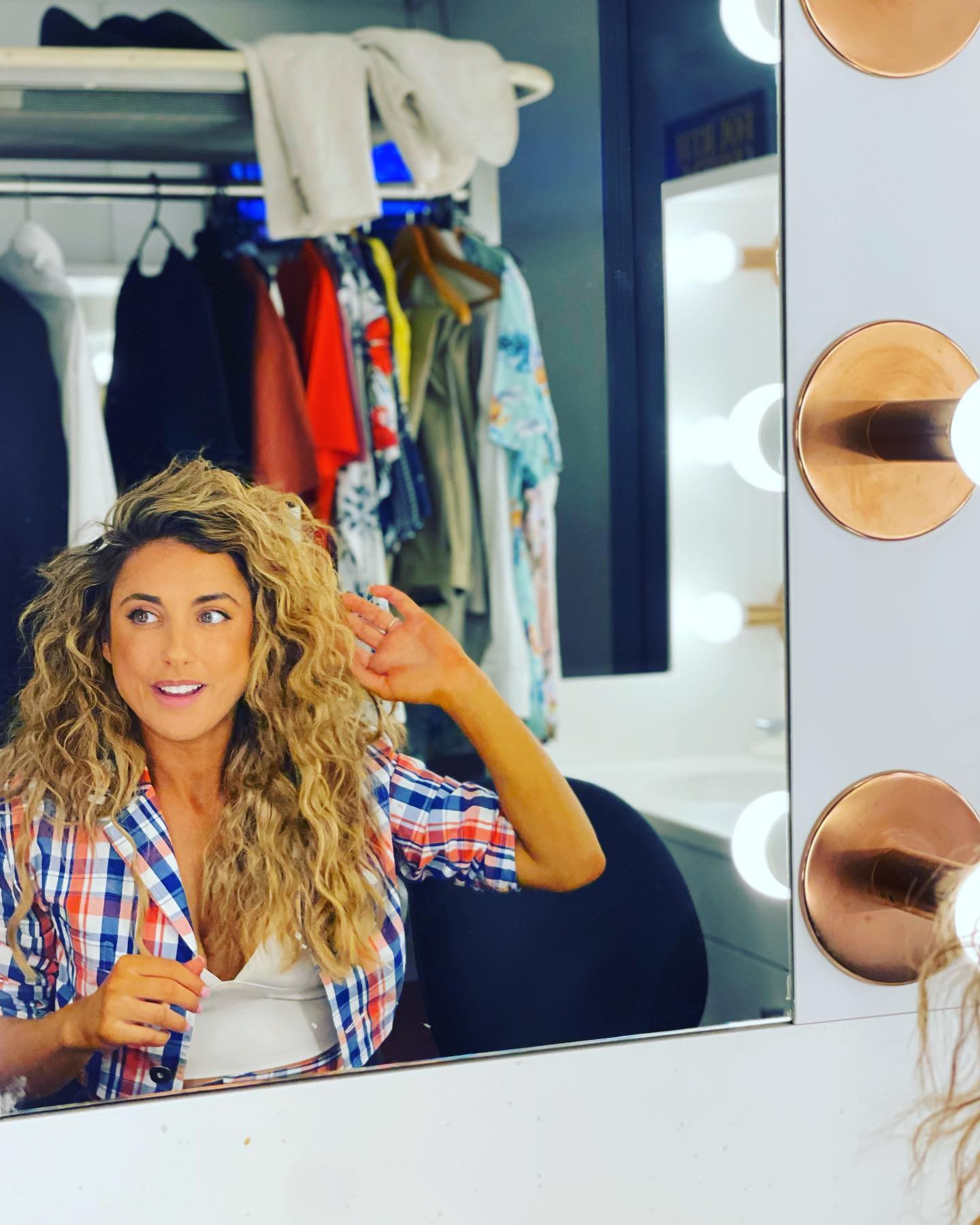Woman with curly hair in a red and blue plaid shirt in front of a mirror with exposed lights, in a dressing room environment.