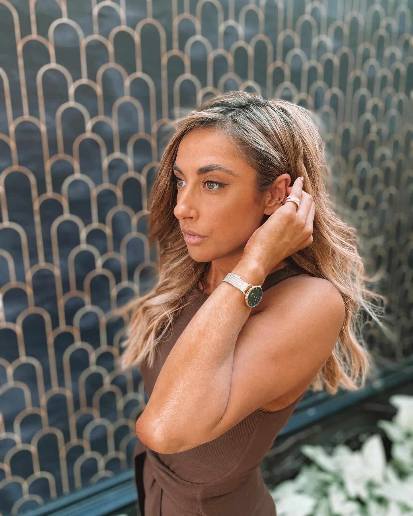 Portrait of a woman with fair skin and long blonde hair, wearing a brown dress and a wristwatch, posing indoors against a patterned wall.