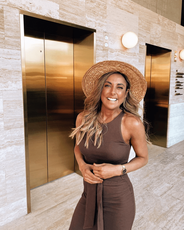A woman in a brown dress and straw hat stands in front of gold elevators in a hotel lobby with patterned marble walls.