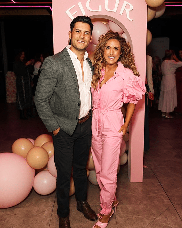A stylish couple poses at an indoor event in front of pink balloons and backdrop. The female is wearing a pink jumpsuit.