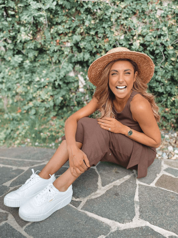 A smiling woman in a brown dress and straw hat sits on stone steps outdoors with lush green foliage as a background, looking cheerful.