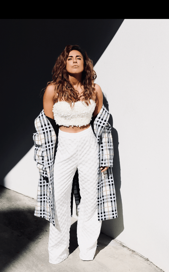A woman poses in the sunlight wearing a white crop top, patterned white pants and a plaid coat against a grey and white wall.