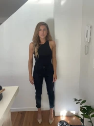 Woman wearing a black sleeveless top and dark jeans standing indoors by a white wall