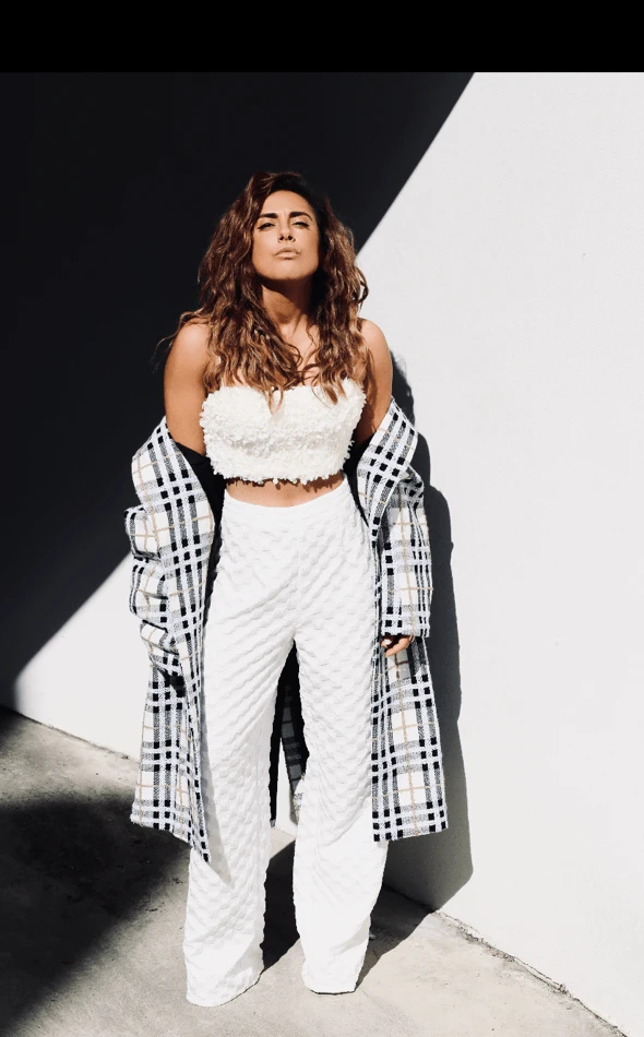 Woman in white strapless crop top and white trousers with a plaid coat draped over her shoulders outdoors.
