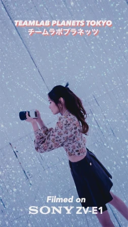 A woman stands in a light installation holding a camera, with soft lighting and a modern aesthetic. The scene is captured at eye level.