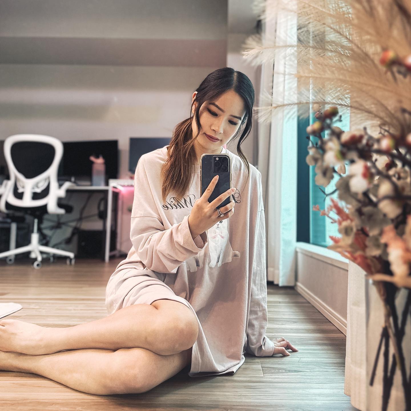 Asian woman in light pink loungewear takes a selfie while sitting cross-legged on a wooden floor in a brightly lit room.