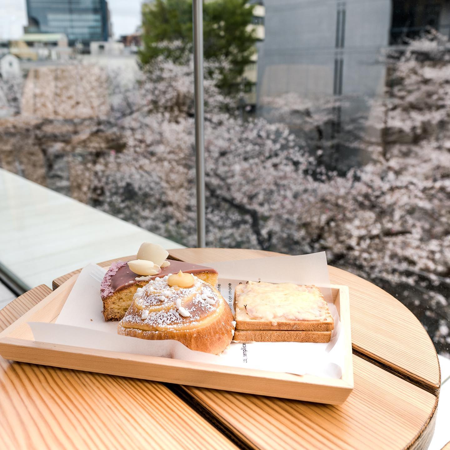 Overhead view of three assorted pastries displayed on a light wood tray, sitting on a wood table indoors with a blurred cityscape backdrop.