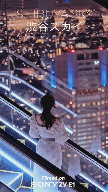 Back view of a woman in a white dress ascending an escalator inside a modern building overlooking the illuminated cityscape at night.