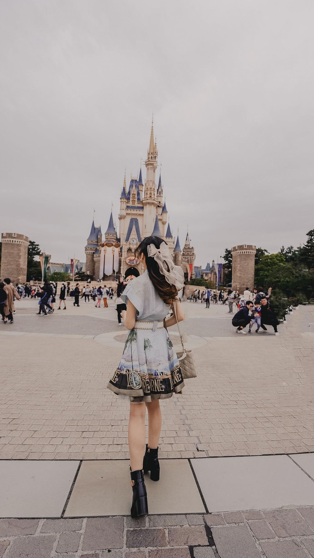 Woman in patterned dress walking towards a fairy tale castle in an amusement park on a cloudy day, carrying a handbag.