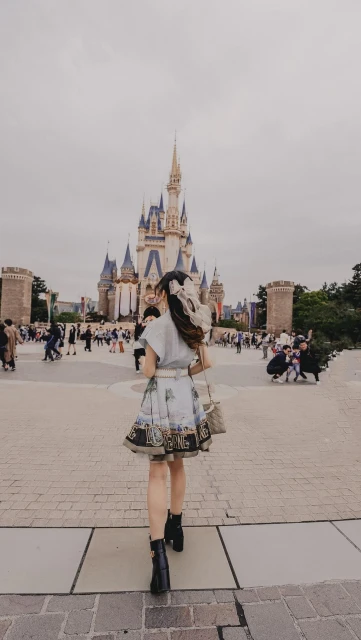 Woman in patterned dress walking towards a fairy tale castle in an amusement park on a cloudy day, carrying a handbag.