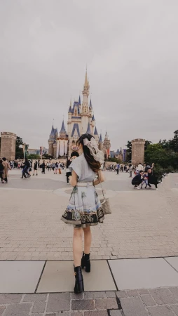 Woman in patterned dress walking towards a fairy tale castle in an amusement park on a cloudy day, carrying a handbag.