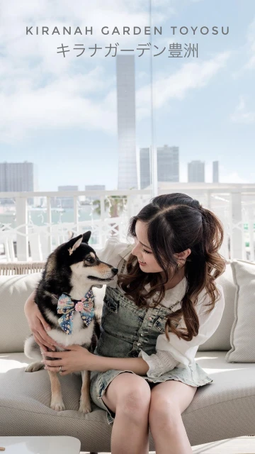 A young adult woman with long brown hair sits on a sofa outside while hugging a Shiba Inu dog wearing a bow tie.