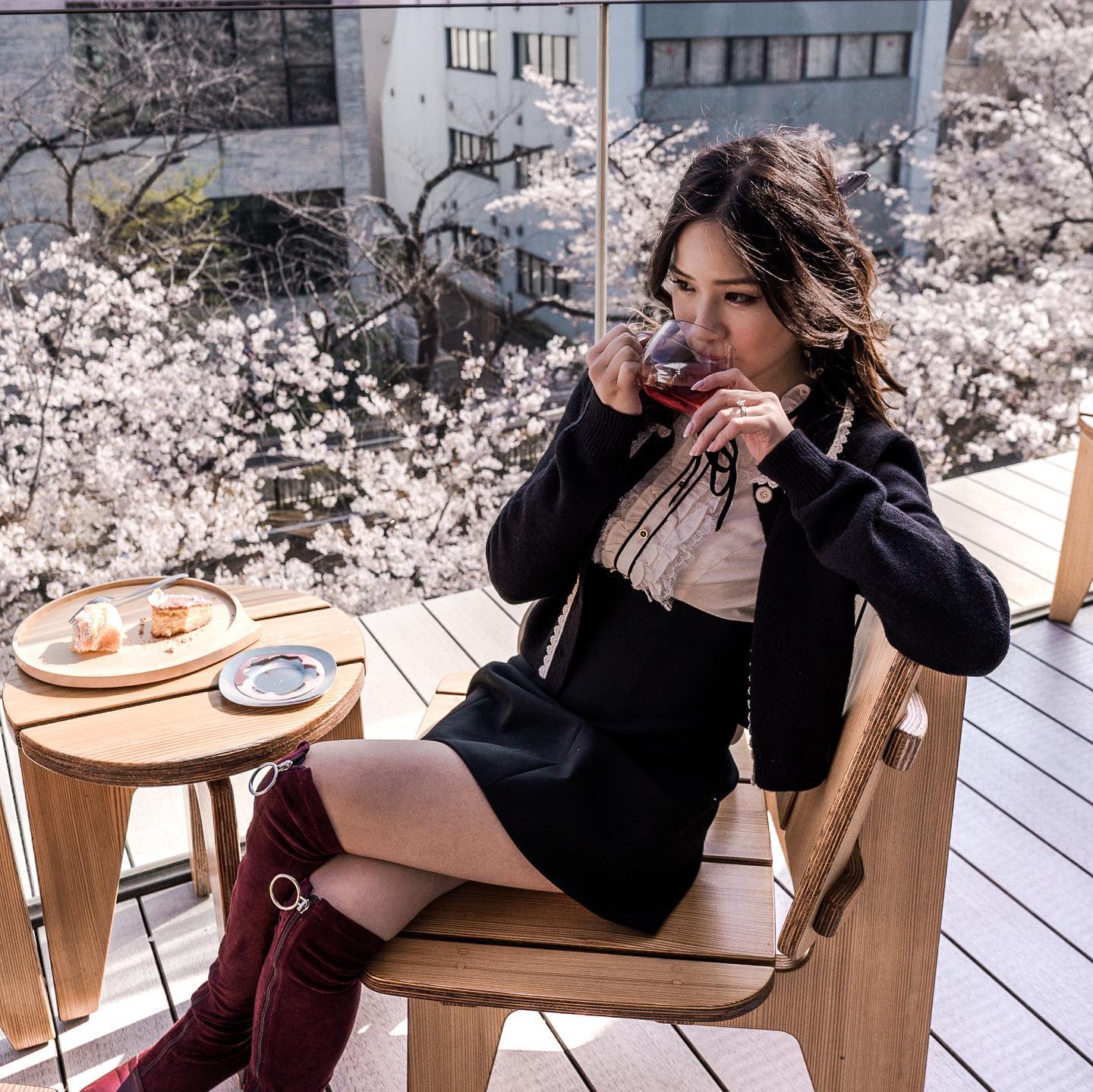 Woman in black dress drinking from a glass on a wooden chair with a table and cherry blossom trees in the background.