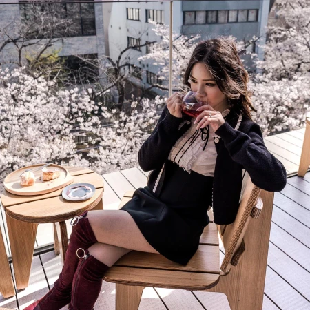 Woman in black dress drinking from a glass on a wooden chair with a table and cherry blossom trees in the background.