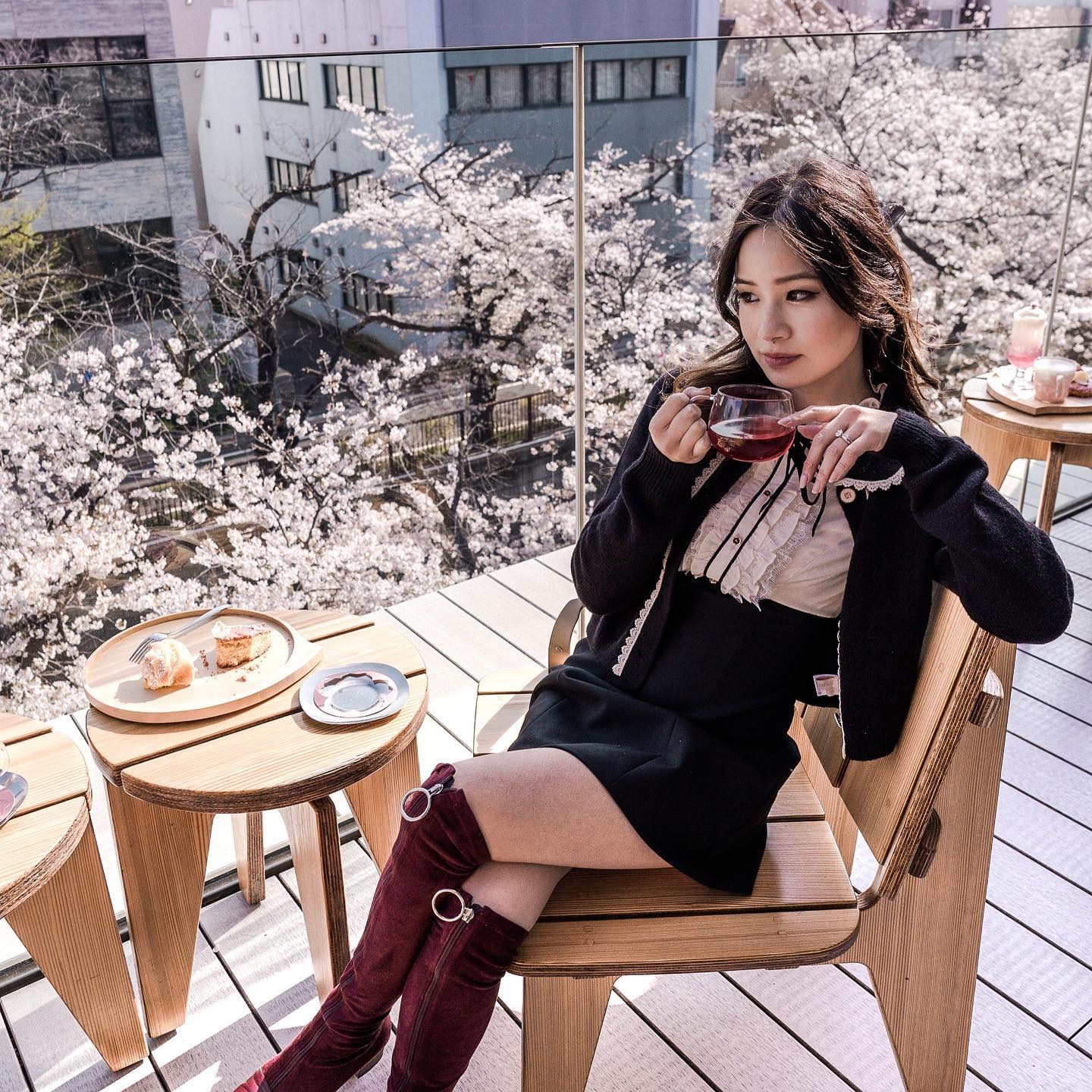 Asian woman in a black dress and burgundy boots sits on a chair on a balcony, holding a glass of tea with cherry blossoms in background.