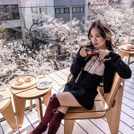 Asian woman in a black dress and burgundy boots sits on a chair on a balcony, holding a glass of tea with cherry blossoms in background.